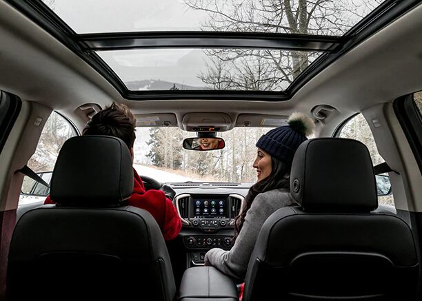 Man and woman in sitting in a car with winter hats and coats on, with the sunroof uncovered to reveal a winter scene