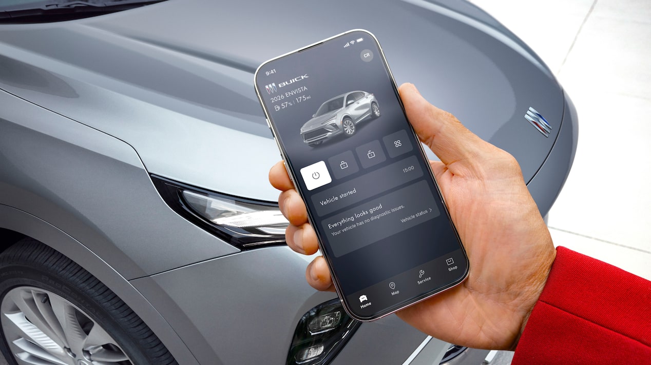 A hand holds a smartphone displaying the Buick mobile app with remote vehicle controls with a grey Envista 2026 in the background of an outdoor setting.