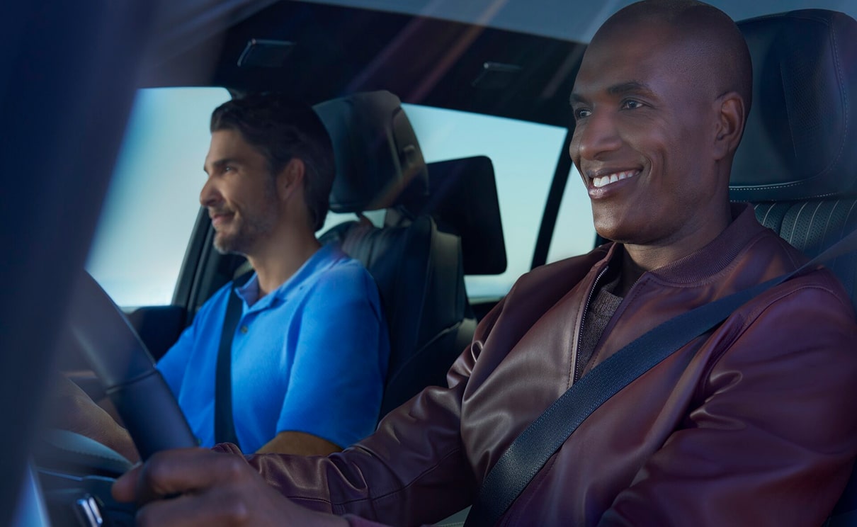 Two men sit in the front seats of a vehicle with their seatbelts on, both smiling as the driver looks ahead at the road.