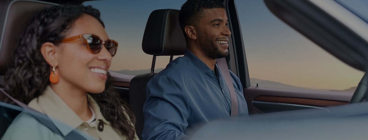 A smiling man and woman sit in the front seats of a vehicle with their seatbelts fastened as they drive during sunset.