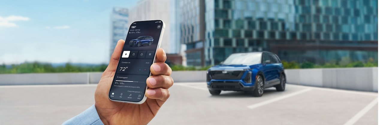 A hand holds a smartphone displaying the myCadillac mobile app with remote vehicle controls while a blue Cadillac VISTIQ is parked in the background of an outdoor rooftop setting.