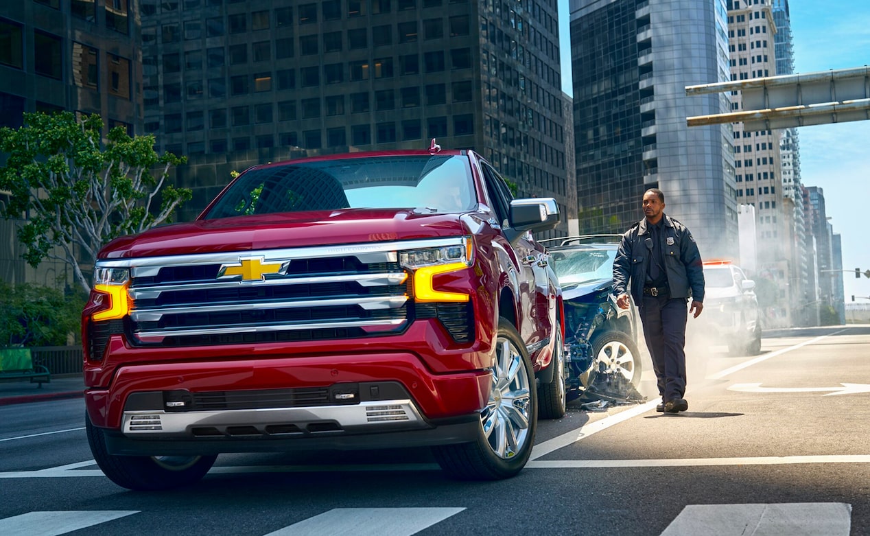 A red Chevrolet Silverado truck is in the foreground of a city street after a minor car accident, with a police officer walking nearby.