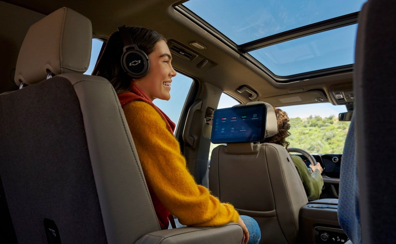 A smiling young woman sits in the back seat of a Chevrolet wearing Chevrolet-branded headphones and looking at a rear-seat entertainment screen that displays app icons like YouTube and Hulu.