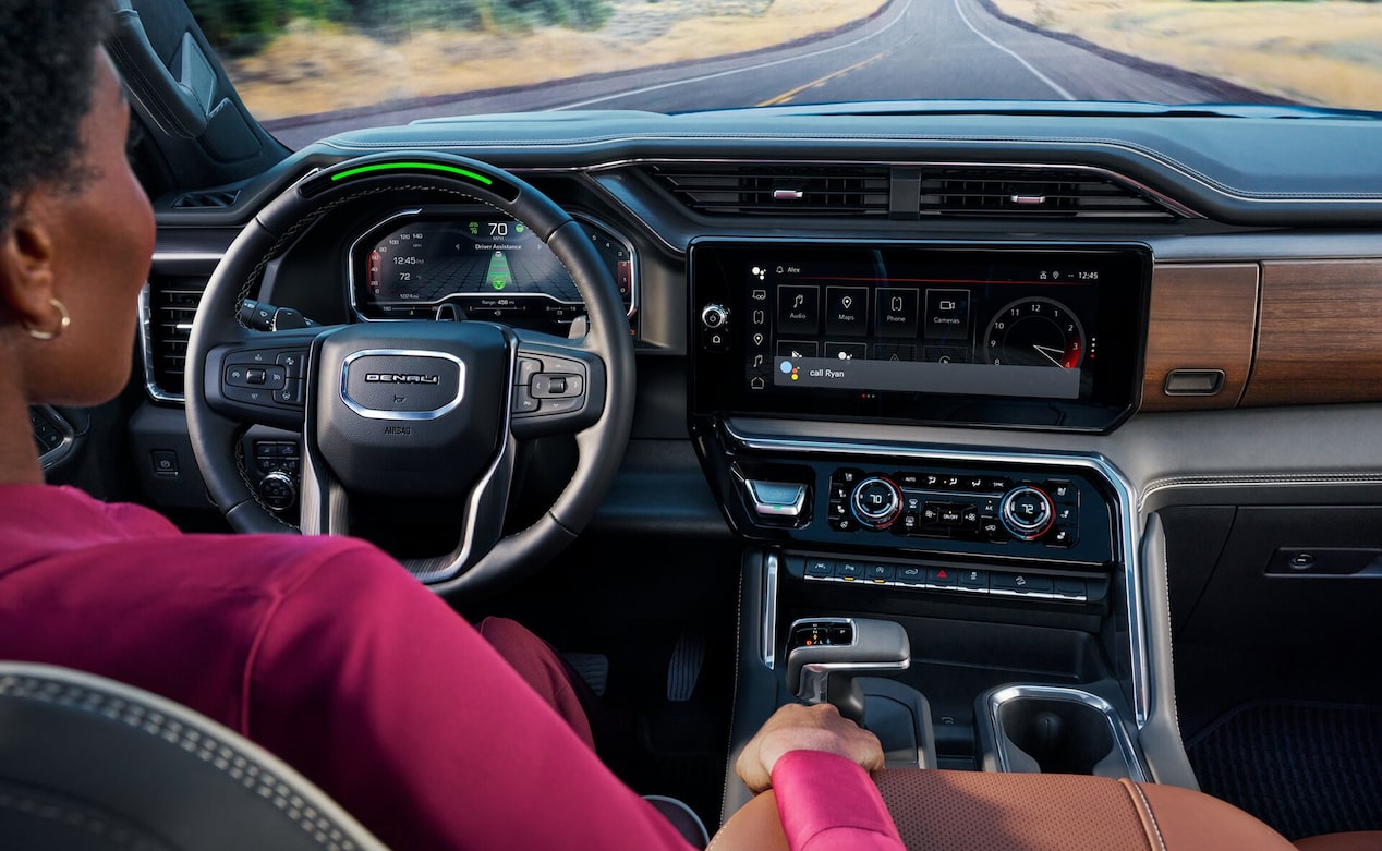 A woman drives a GMC Denali with her hands near the steering wheel while the infotainment screen displays Google built-in features and a voice assistant prompt.