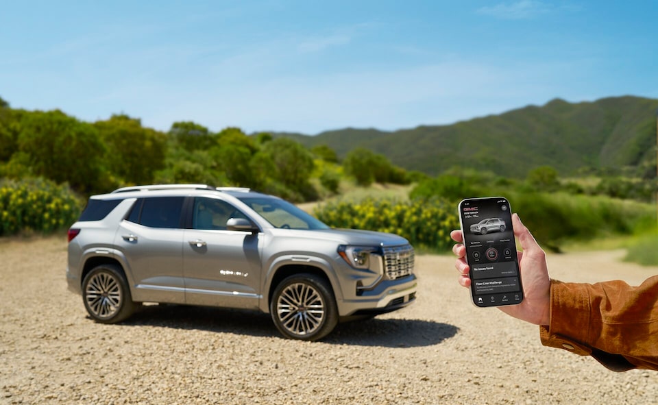 A person holds a smartphone displaying the myGMC mobile app with remote start and lock features in front of a silver GMC Terrain parked in a rural, hilly landscape.