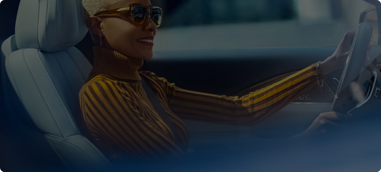 A smiling woman with short blonde hair and sunglasses is driving a car.