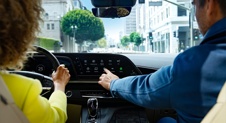 A woman in a yellow jacket holding the steering wheel and a man in a blue jacket pressing a button on the infotainment system